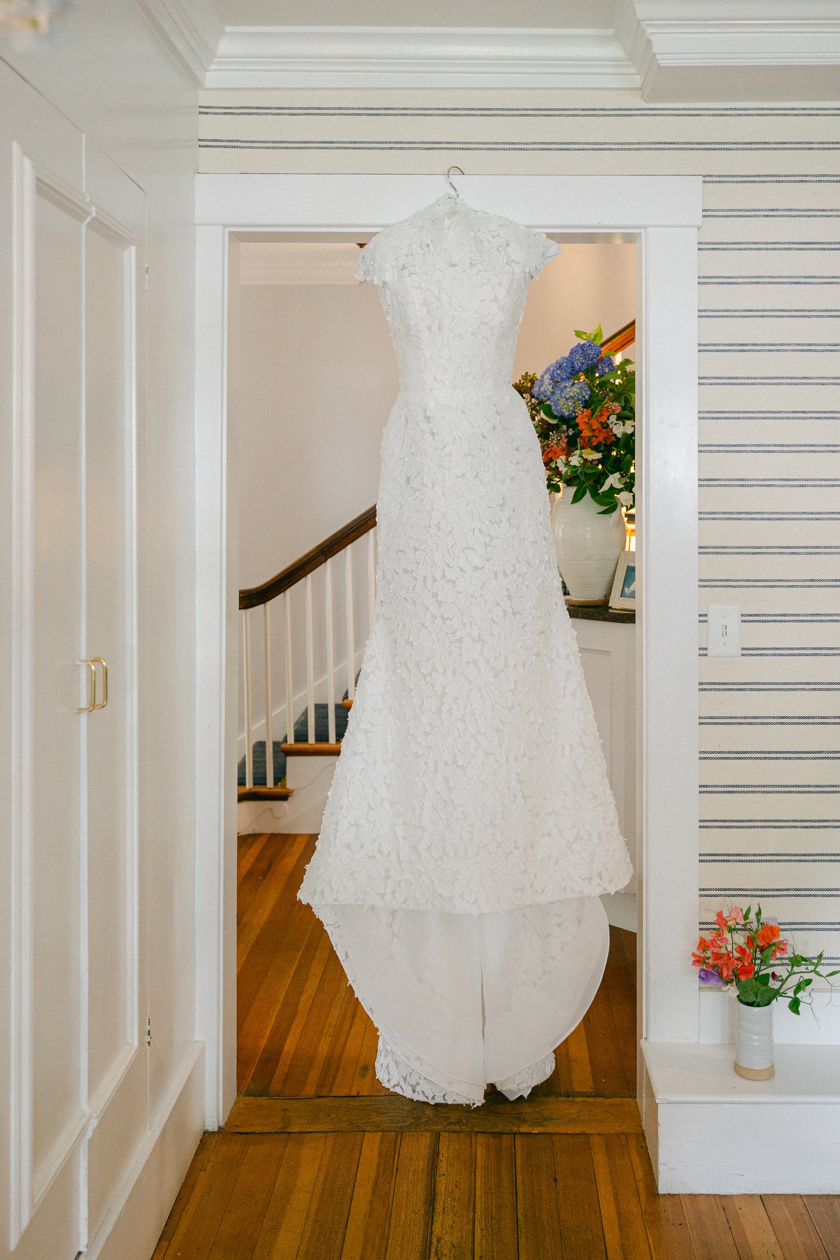 A white wedding dress with lace details hanging in a doorway with striped wallpaper, a vase with colorful flowers on the side table, and hardwood floors at Lambert's Cove Inn on Martha's Vineyard.