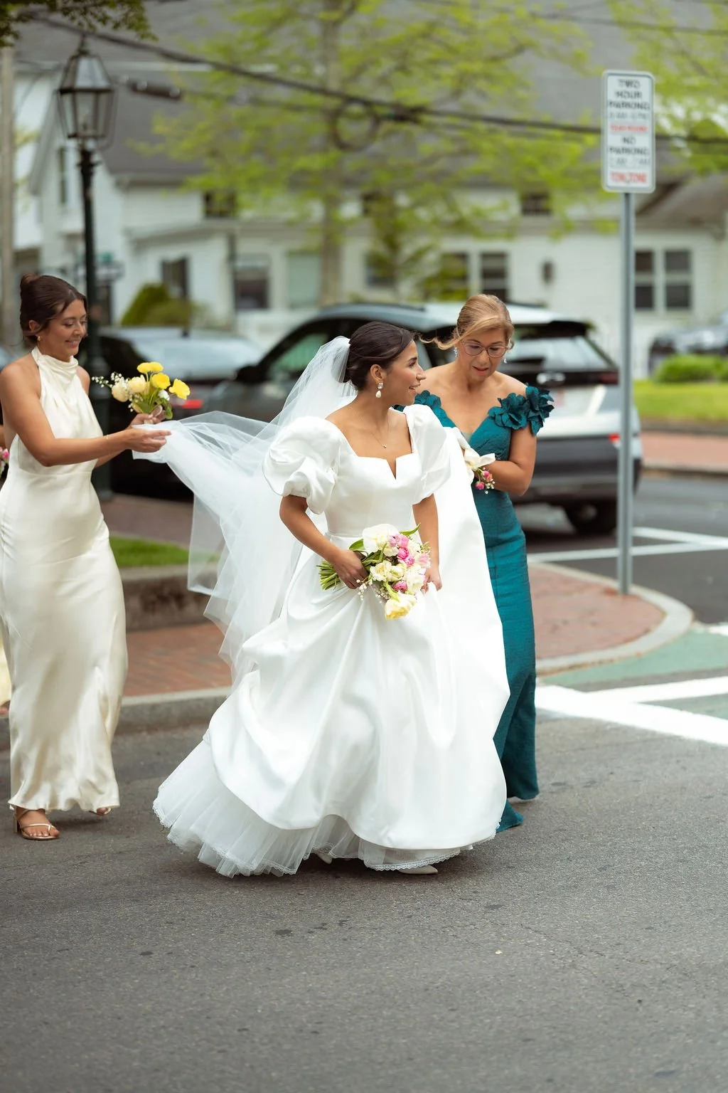 Bride in a white wedding dress holding a bouquet, walking outside with bridesmaids dressed in elegant gowns, one with a green dress and boutonniere, at Lambert's Cove Inn on Martha's Vineyard.