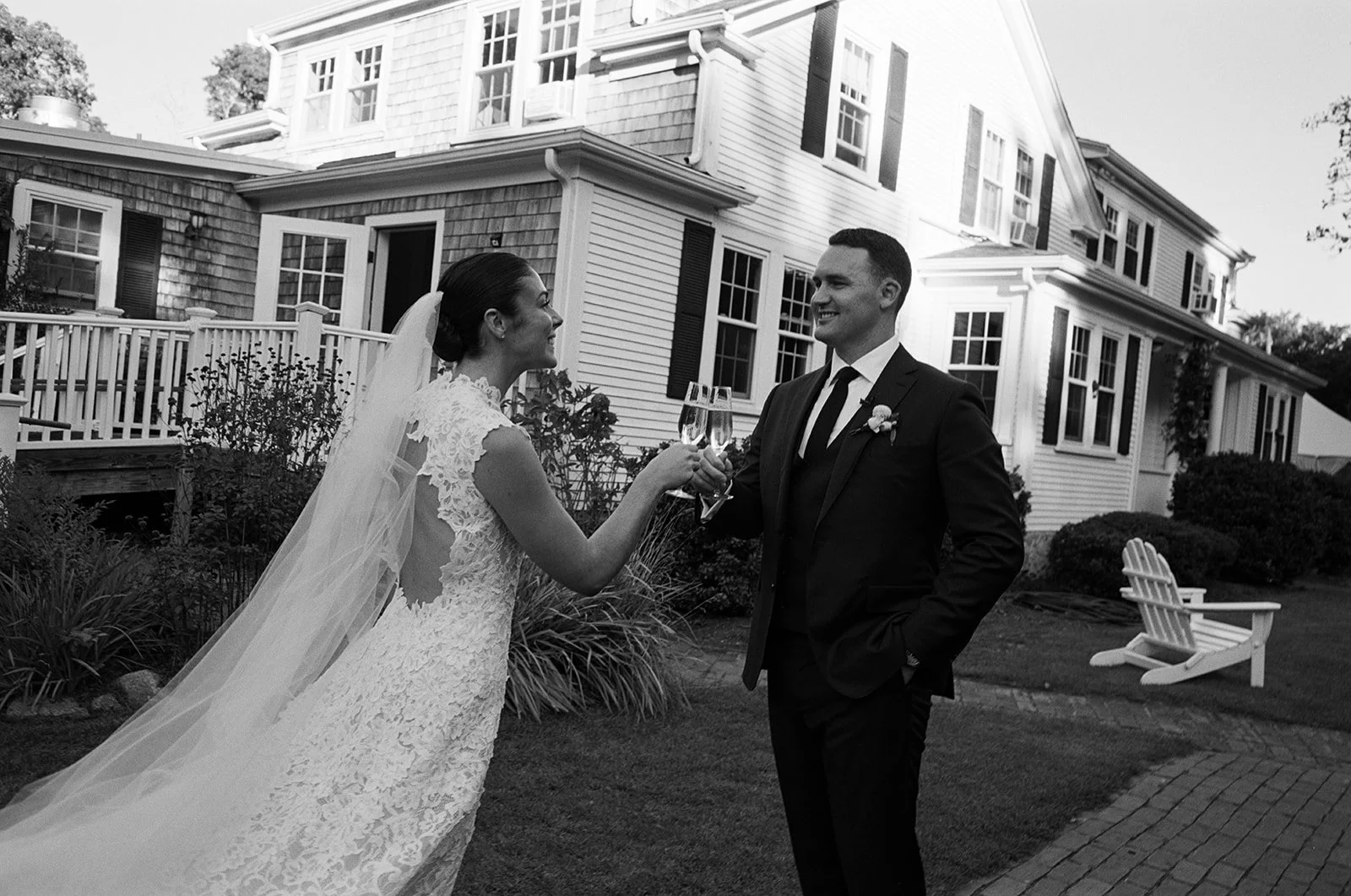 Black and white photo of a newlywed couple celebrating with champagne outside Lambert's Cove Inn. The bride is wearing a lace wedding gown and the groom is in a suit. They are smiling at each other while holding glasses of champagne.