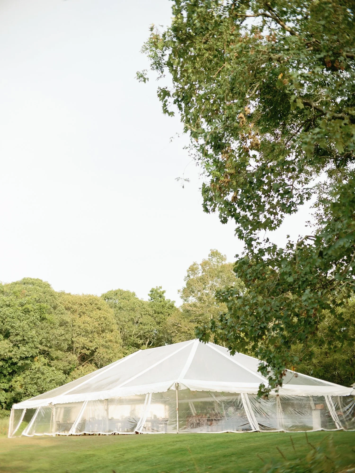 A large white wedding tent set up outdoors on a grassy area at Lambert's Cove Inn on Martha's Vineyard, surrounded by trees with green foliage, under a clear sky.