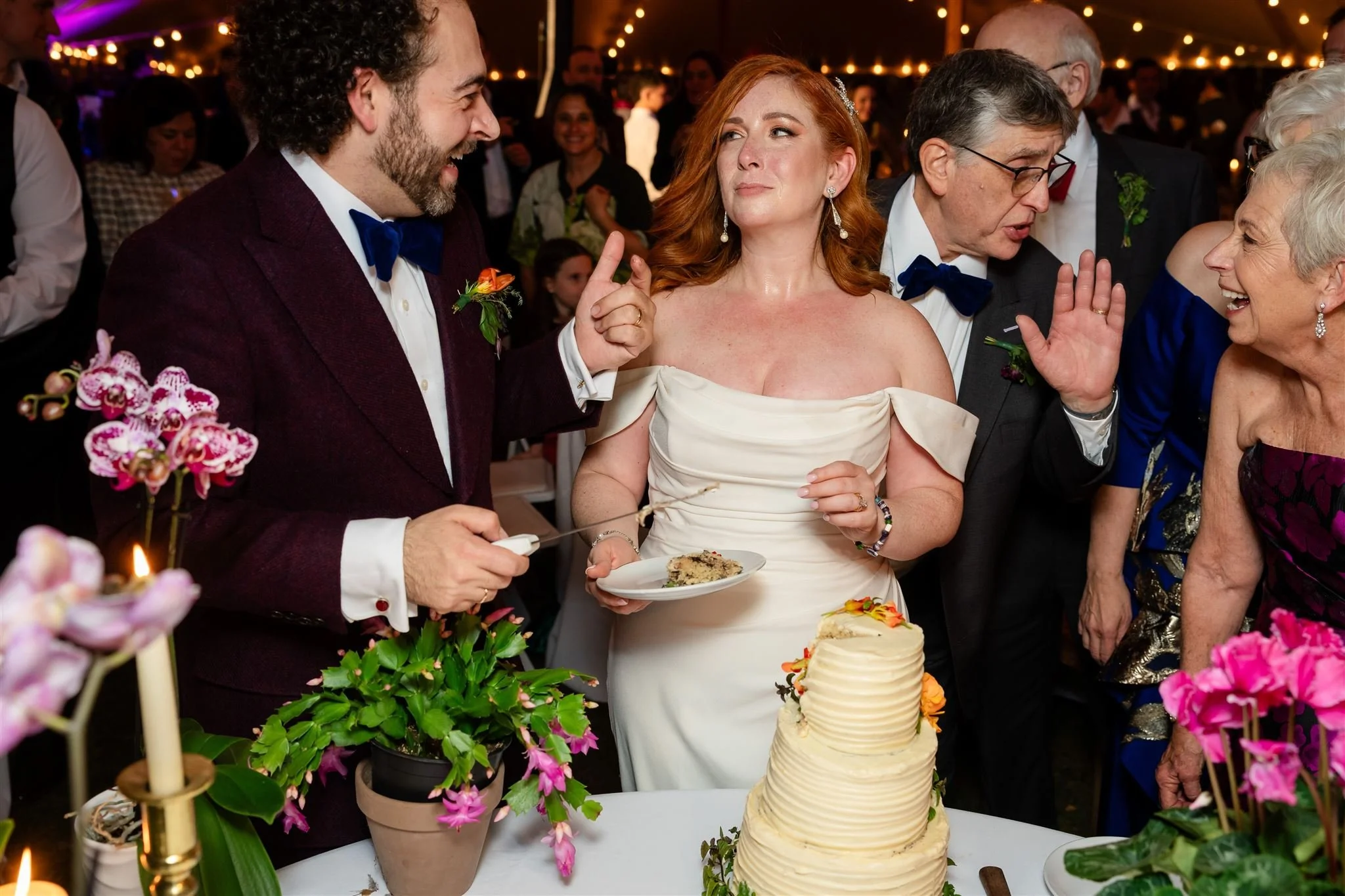 A wedding reception scene with a bride and groom cutting a tiered wedding cake, surrounded by floral decorations and guests. The groom is wearing a dark suit with a blue bow tie, and the bride is in a white off-shoulder dress. The background features