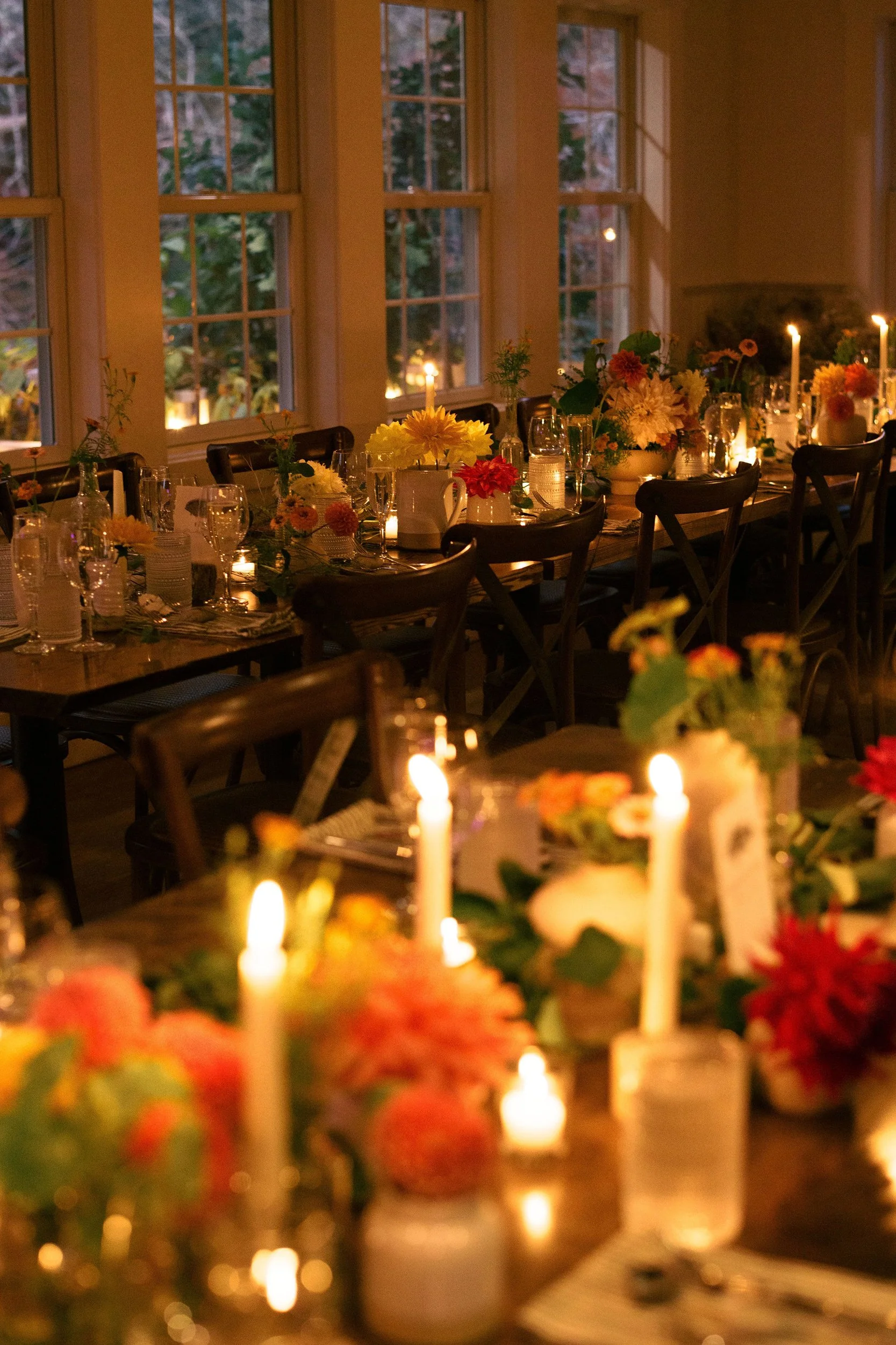 A candlelit dinner table decorated with flowers and candles, set near large windows showing the outdoors.