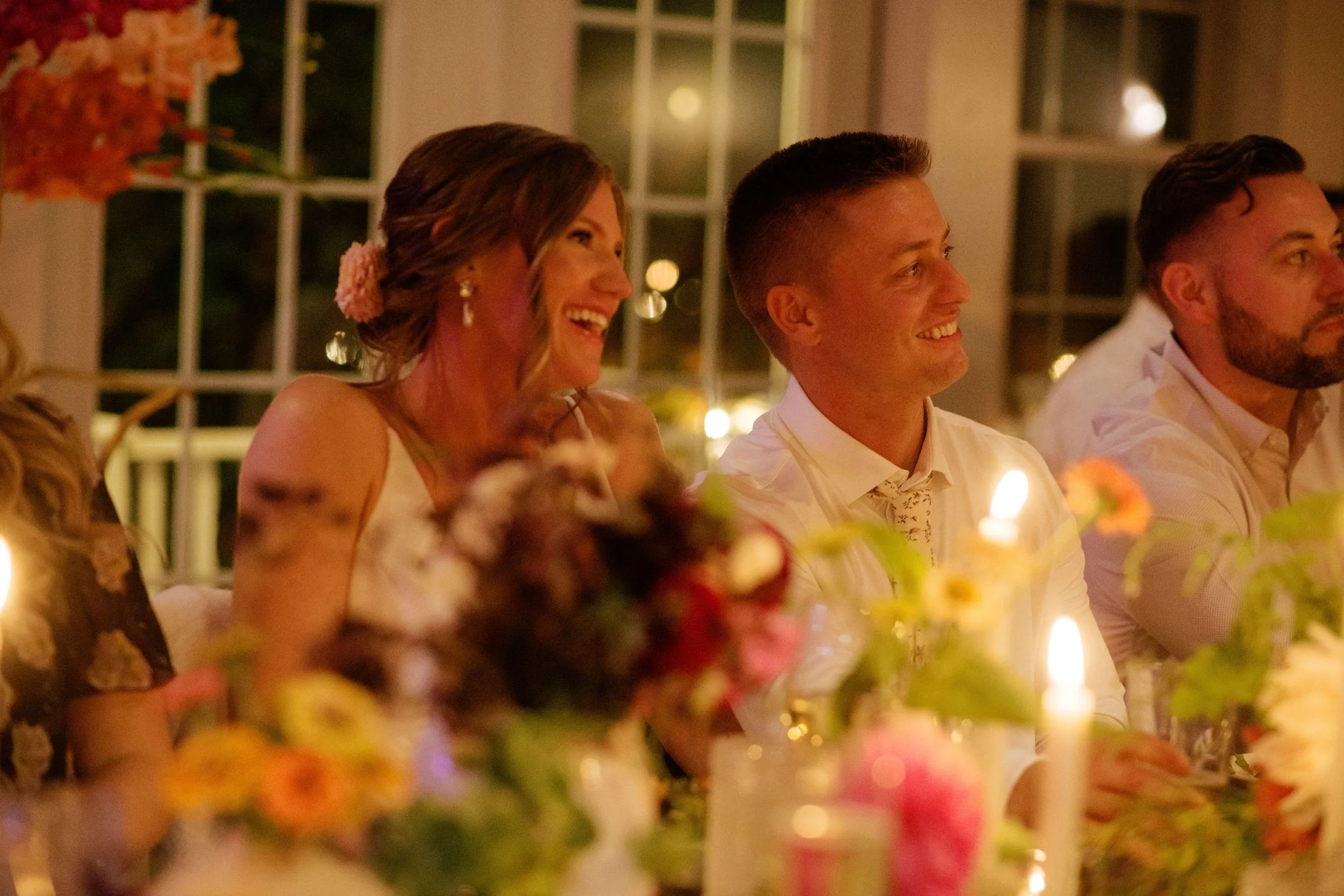 People sitting at a table during a wedding reception at Lambert's Cove Inn on Martha's Vineyard, smiling and enjoying the moment, with candles and floral decorations in a warmly lit room.