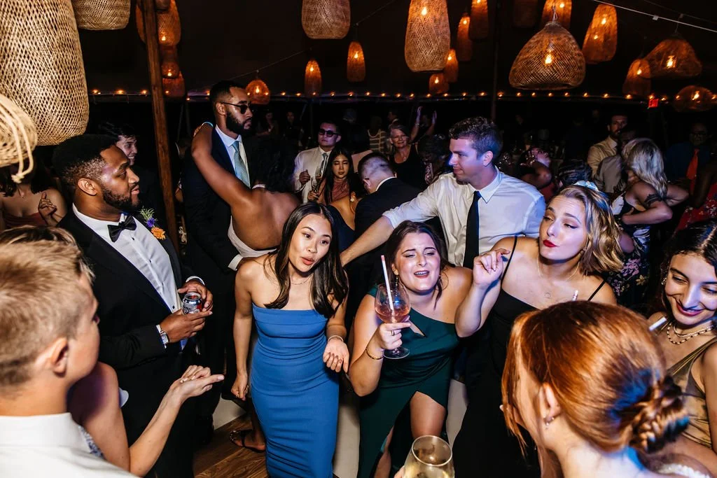 People dancing and socializing at a lively indoor party with hanging wicker light fixtures during a wedding reception at Lambert's Cove Inn on Martha's Vineyard.