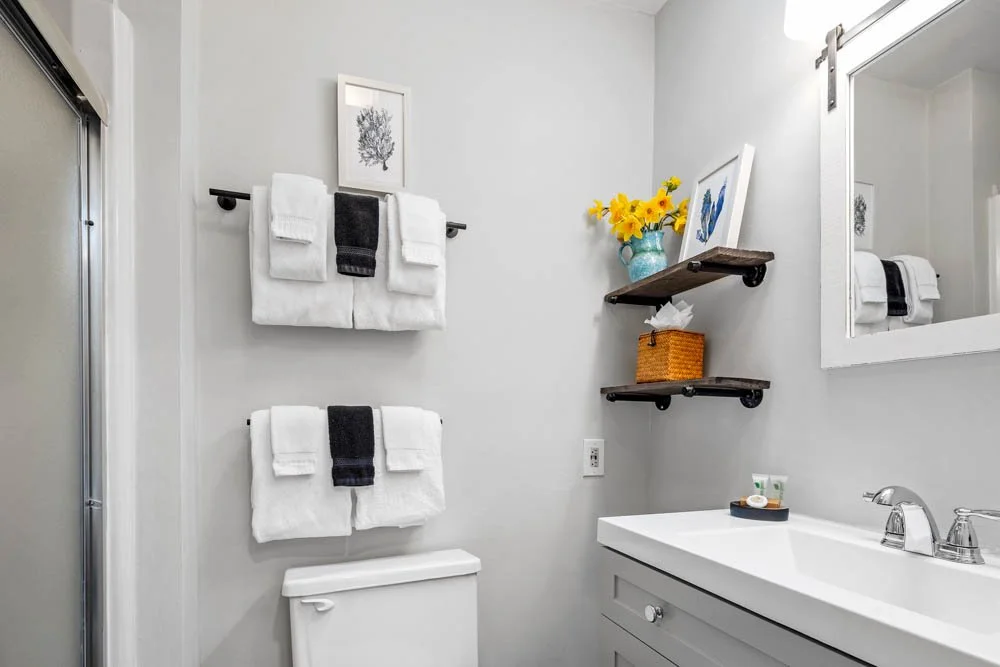 Bathroom with white towels, black towels, a framed coral print, shelves with a vase of yellow flowers, a wicker basket, and a mirror above a white sink.