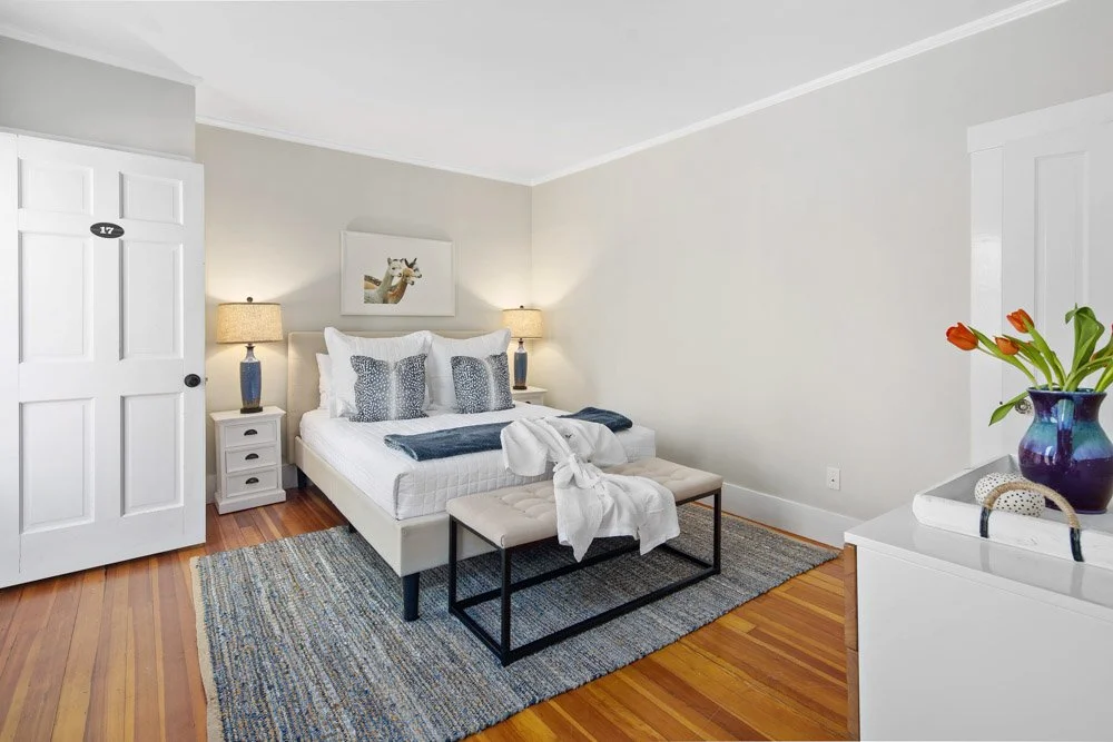 A tidy bedroom with a white bed, three decorative pillows, two bedside lamps, a small framed art piece, a bench at the foot of the bed with a blanket, part of a white dresser with a purple vase with orange flowers, and a wooden floor with a multicolo