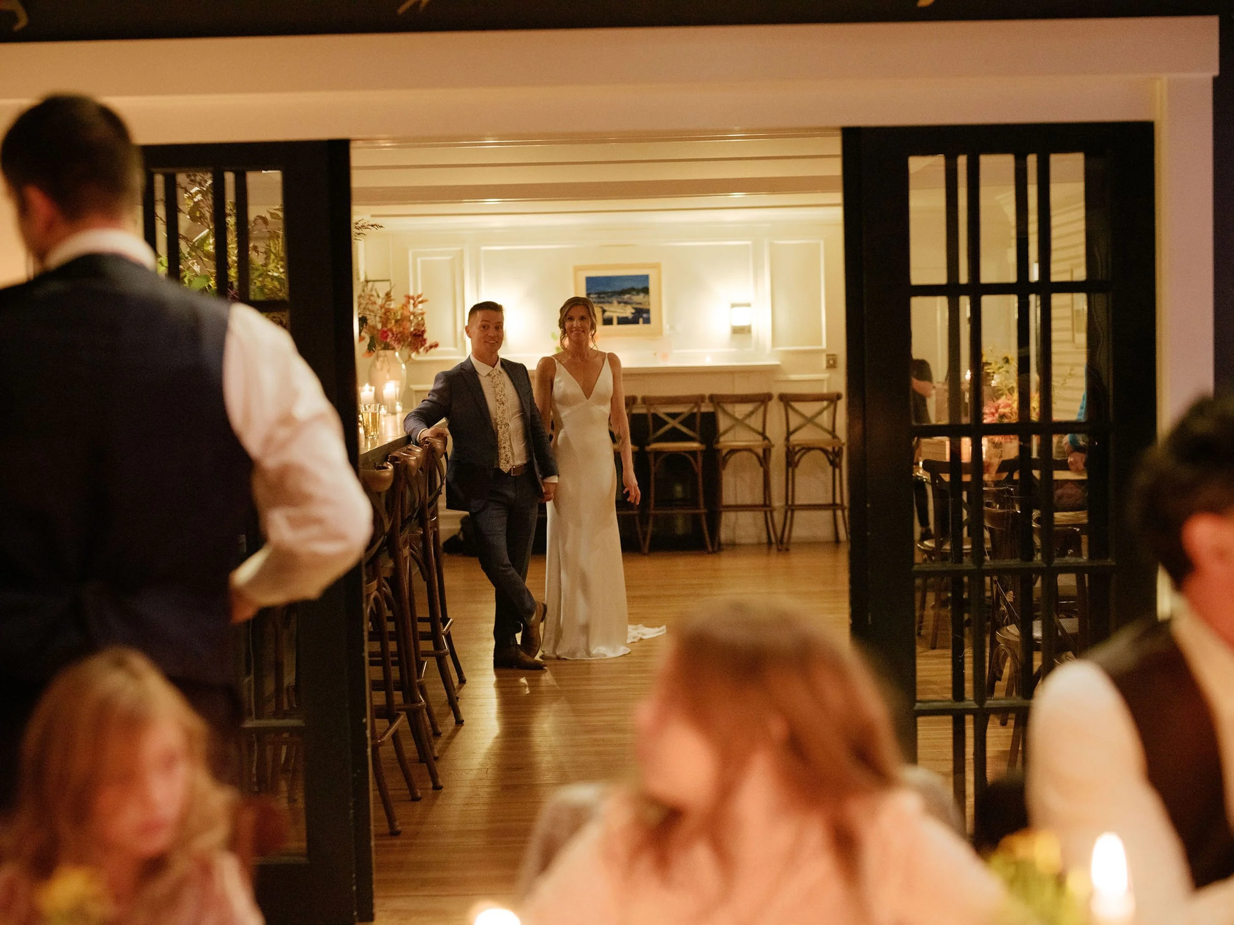 A bride and groom stand at a wedding reception at Lambert's Cove Inn on Martha's Vineyard, smiling and holding hands, with guests in the background inside a warmly lit room.