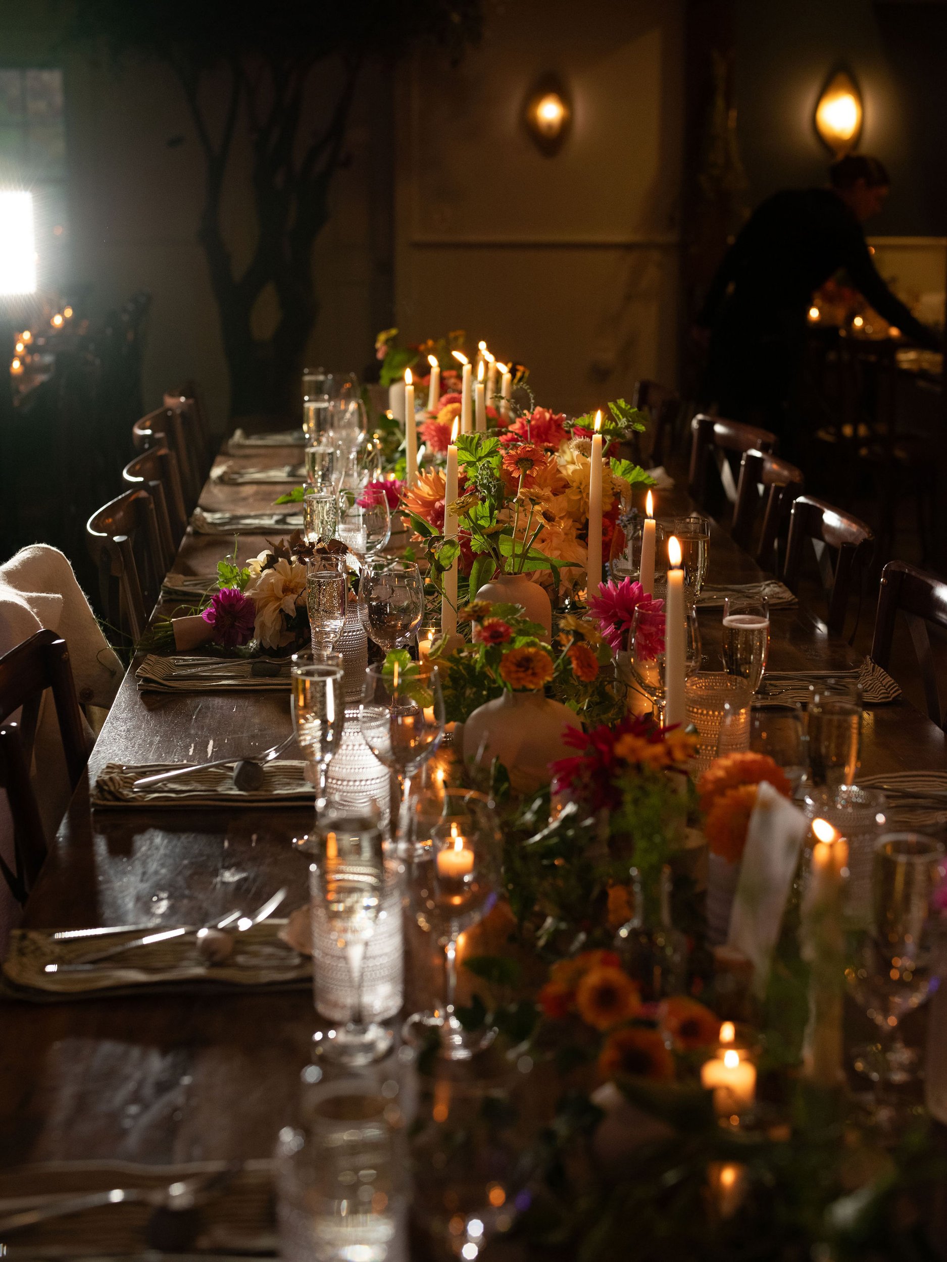 A long dining table set for a special occasion with books of flowers and candles, in a dimly lit room with wall sconces and a person organizing items in the background.