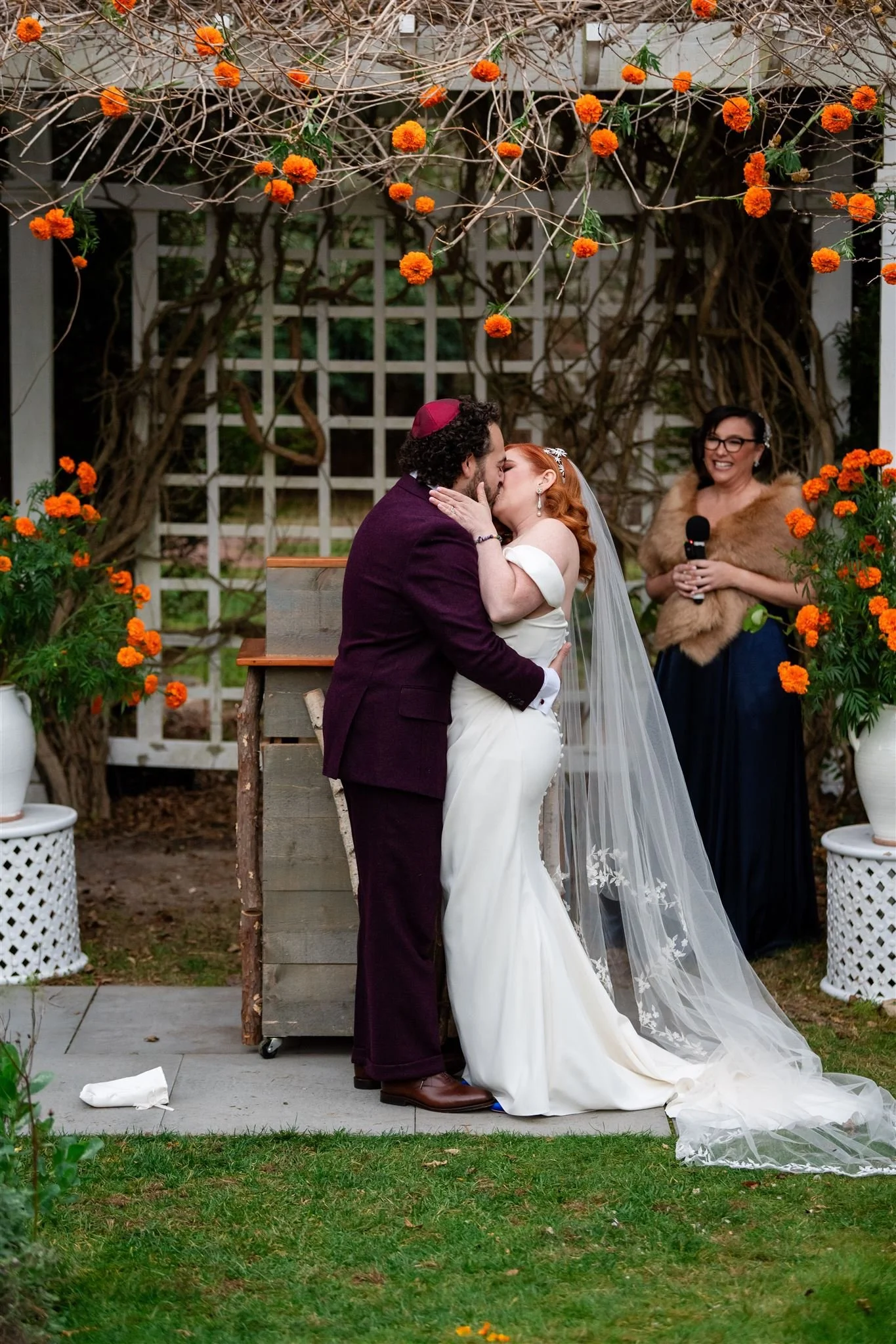 A couple is sharing a kiss during their wedding ceremony in an outdoor garden setting. The bride is in a white wedding gown with a long veil, and the groom is wearing a dark suit and a pink kippah. An officiant holding a microphone stands nearby, smi