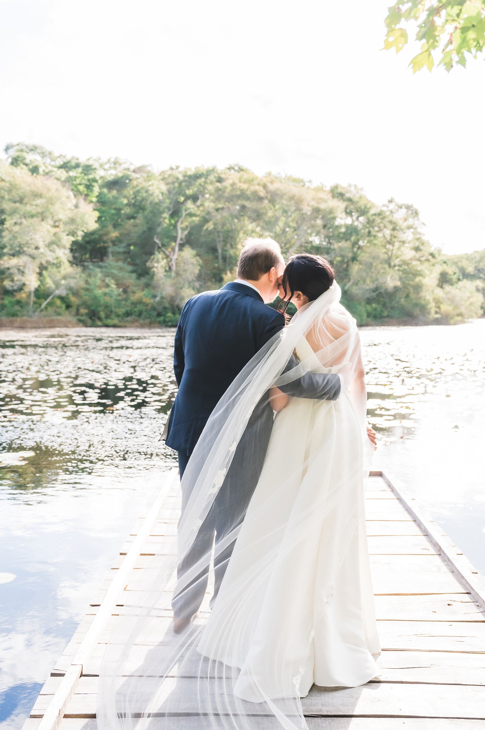 A bride and groom sharing a kiss on a wooden dock by a body of water, with trees in the background at Lambert's Cove Inn on Martha's Vineyard.