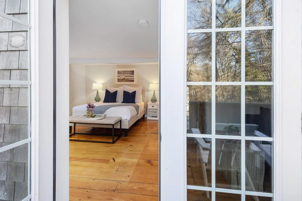 View through a glass door into a bedroom with a bed, side tables with lamps, artwork on the wall, and a wooden floor.