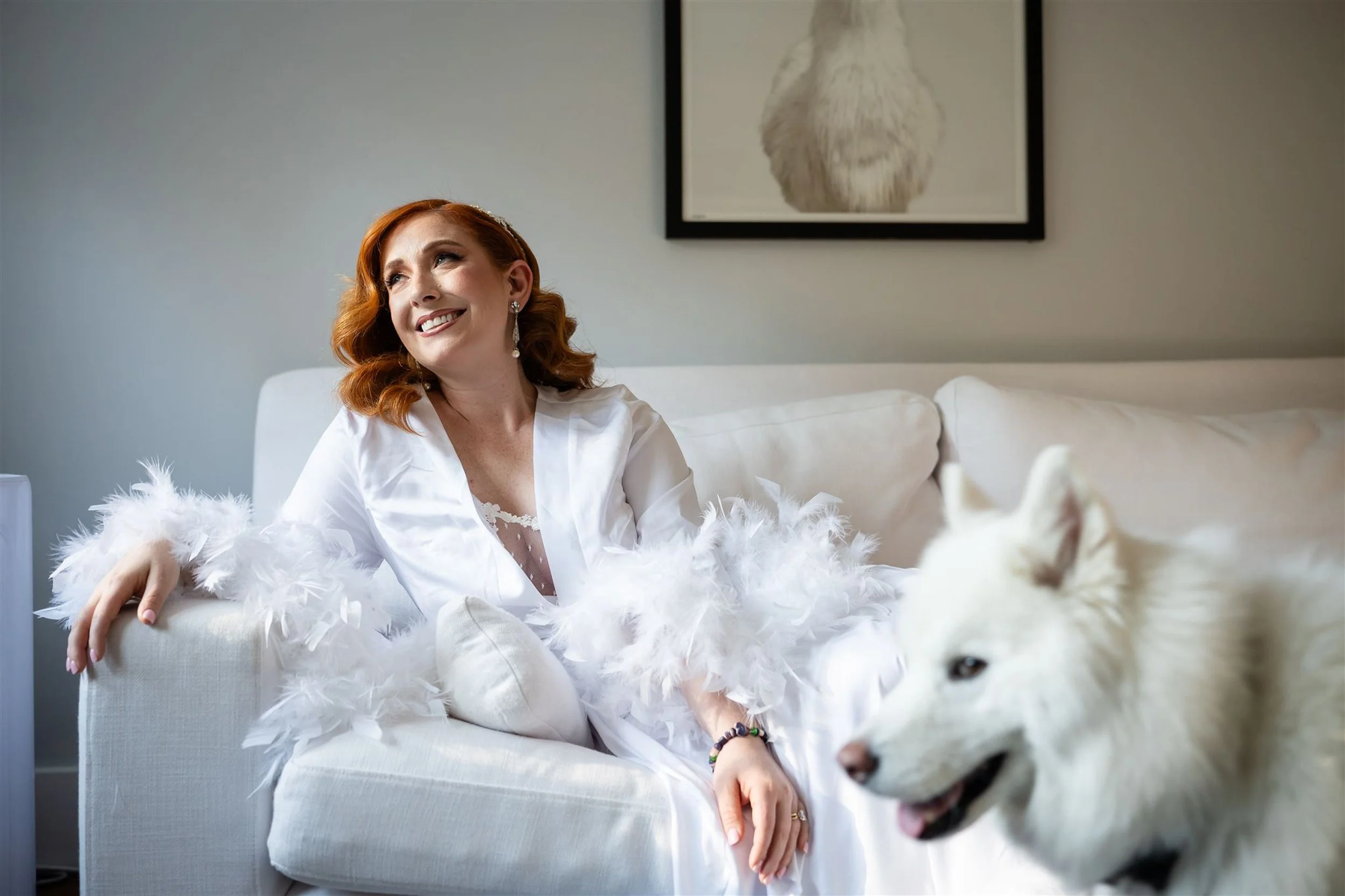A woman with red hair, wearing elegant earrings and a white robe with feather accents, sitting on a white sofa in a modern room with a framed animal picture on the wall and a white dog nearby.