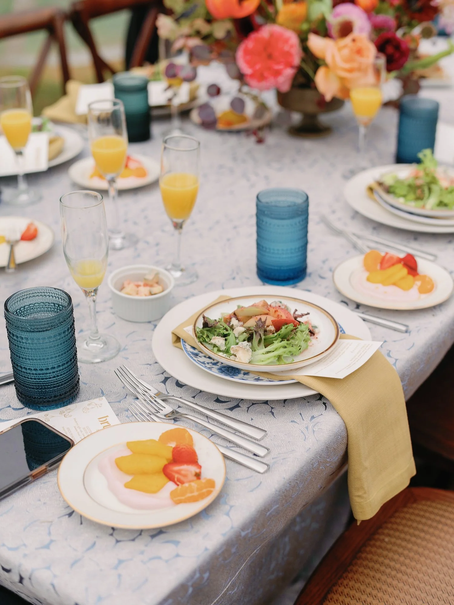 Table set for a meal with plates of salad and tropical fruit, glasses of orange juice, and blue glasses, on a white tablecloth with a yellow napkin, with a large floral centerpiece in the background at Lambert's Cove Inn on Martha's Vineyard.