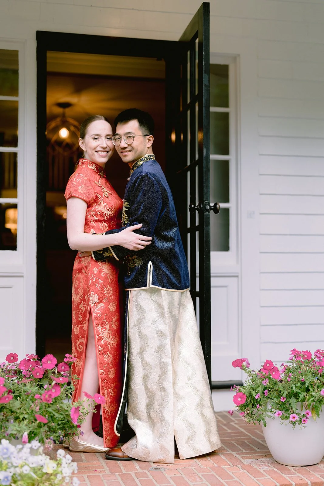 A smiling couple hugging each other at the doorway of Lambert's Cove Inn on Martha's Vineyard, dressed in traditional Asian attire, with pink flowers in pots on brick ground outside.
