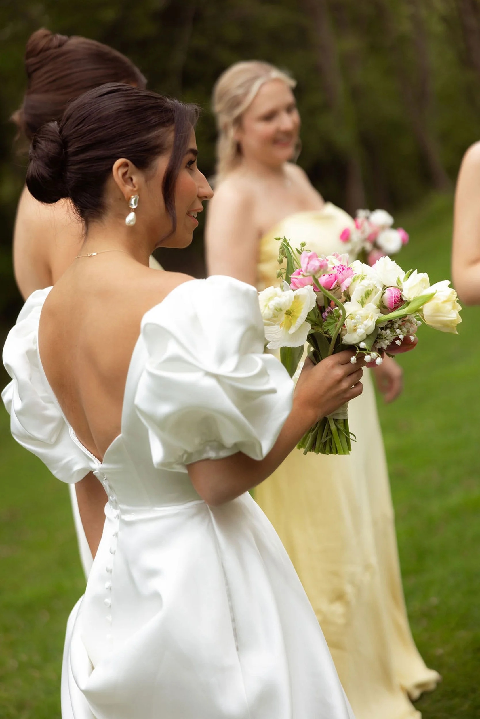 A woman in a white dress is holding a bouquet of flowers at an outdoor wedding reception at Lambert's Cove Inn on Martha's Vineyard, with other women in formal attire nearby and trees in the background.