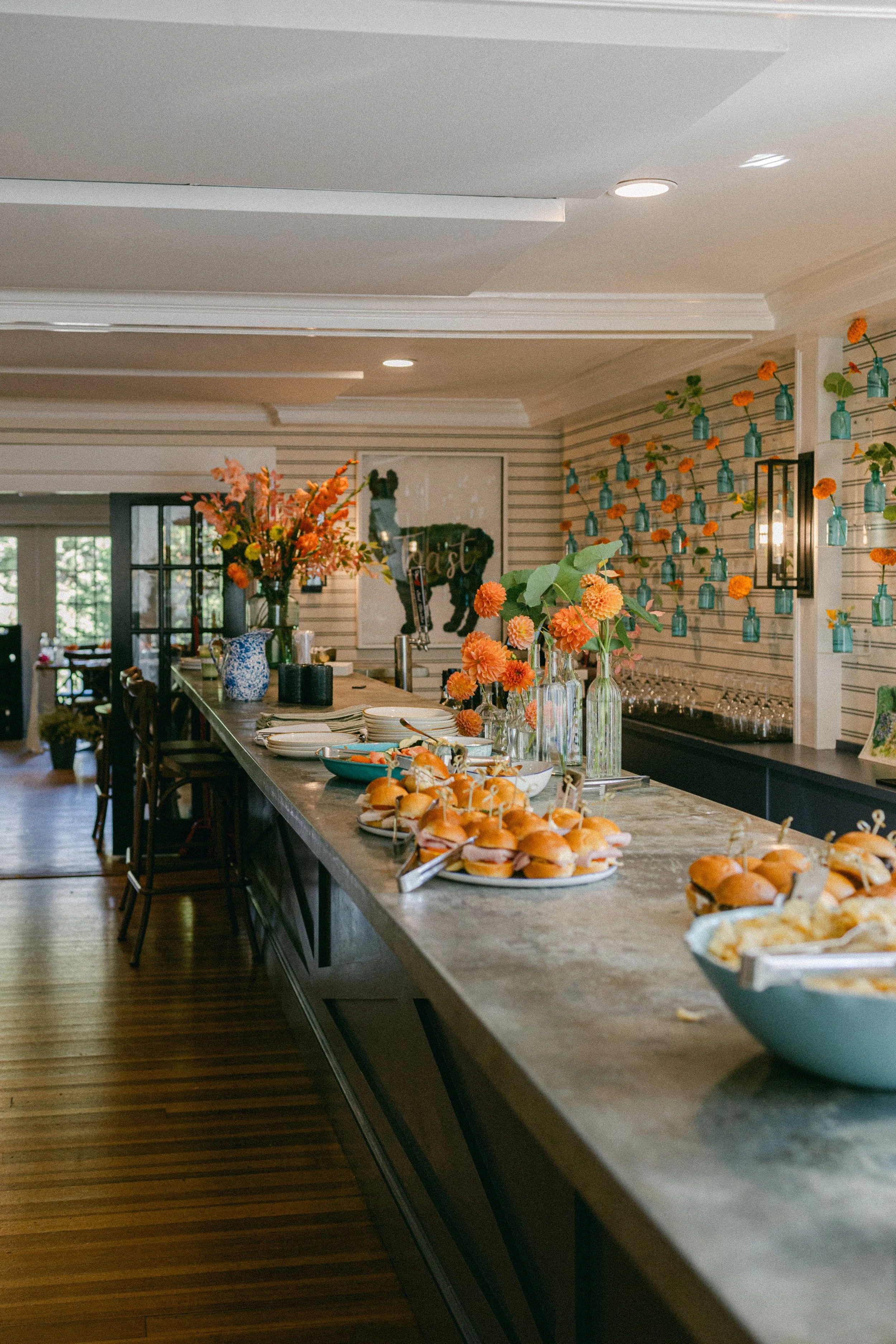 A buffet table with small sandwiches and pastries, decorated with orange and peach flowers in vases, in a well-lit room with floral wall decor and blue bottles hanging on the wall.