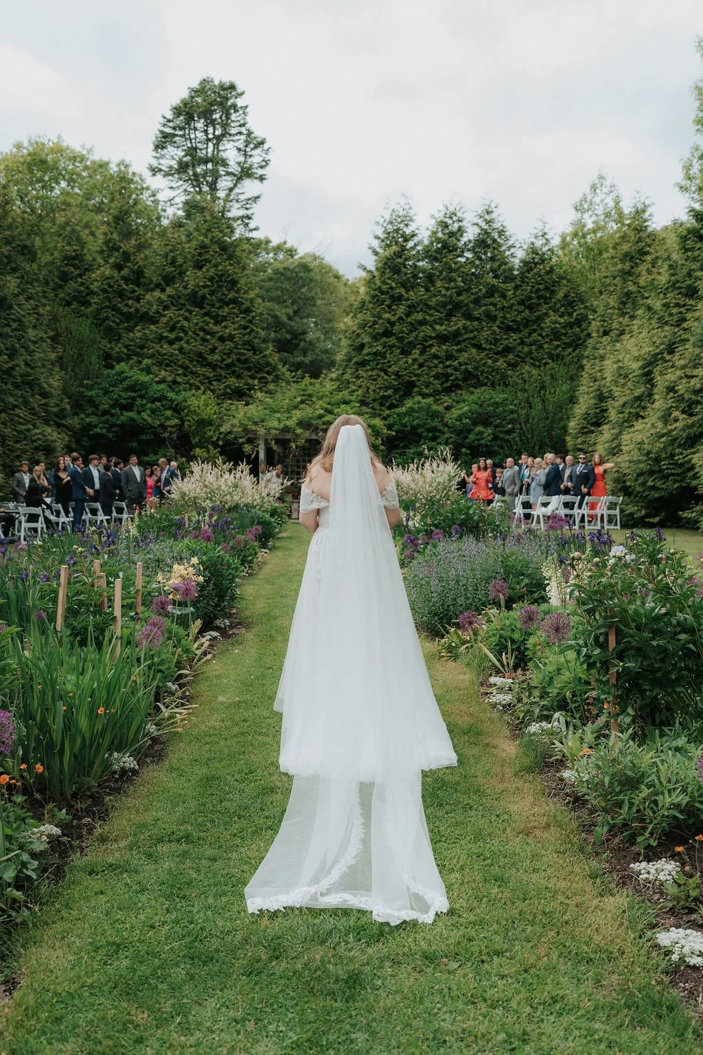 Bride with a veil walking down an aisle during a wedding ceremony at Lambert's Cove Inn on Martha's Vineyard, with guests seated on both sides against lush trees.