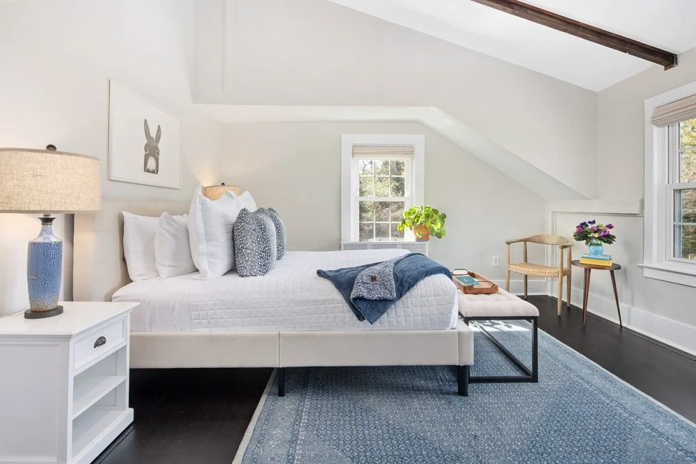 A bright bedroom with white walls, a bed with white bedding, gray pillows, and a blue throw blanket. There is a white nightstand with a textured blue table lamp on the left. A small window behind the bed shows greenery outside. A wooden chair, side t