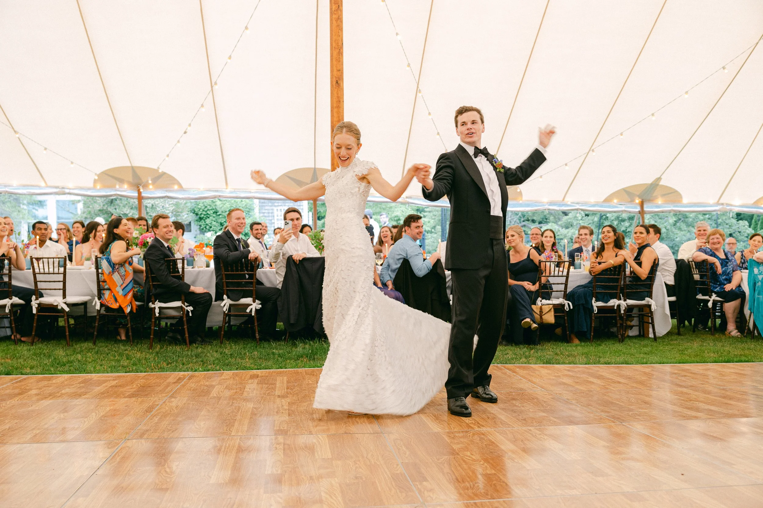 A bride and groom dancing at their wedding reception inside a large tent with string lights. The bride is in a white lace wedding gown, and the groom is in a tuxedo. They are holding hands and smiling, while guests sit and watch the celebration.