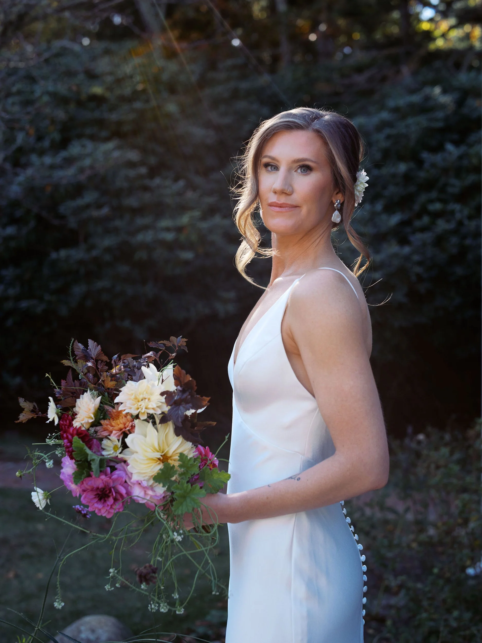 A woman in a white dress holding a colorful bouquet of flowers outdoors at sunset during a wedding at Lambert's Cove Inn on Martha's Vineyard with greenery in the background.