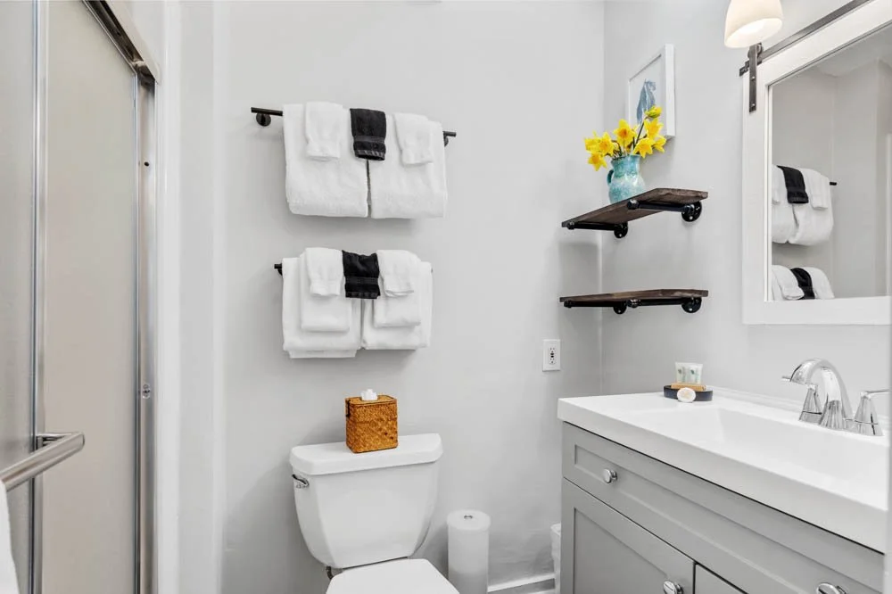 A modern bathroom featuring a toilet with a woven basket on top, white and black towels on wall-mounted racks, a white vanity with a sink, a large mirror, floating shelves with a vase of yellow flowers, and a shower enclosure on the left.