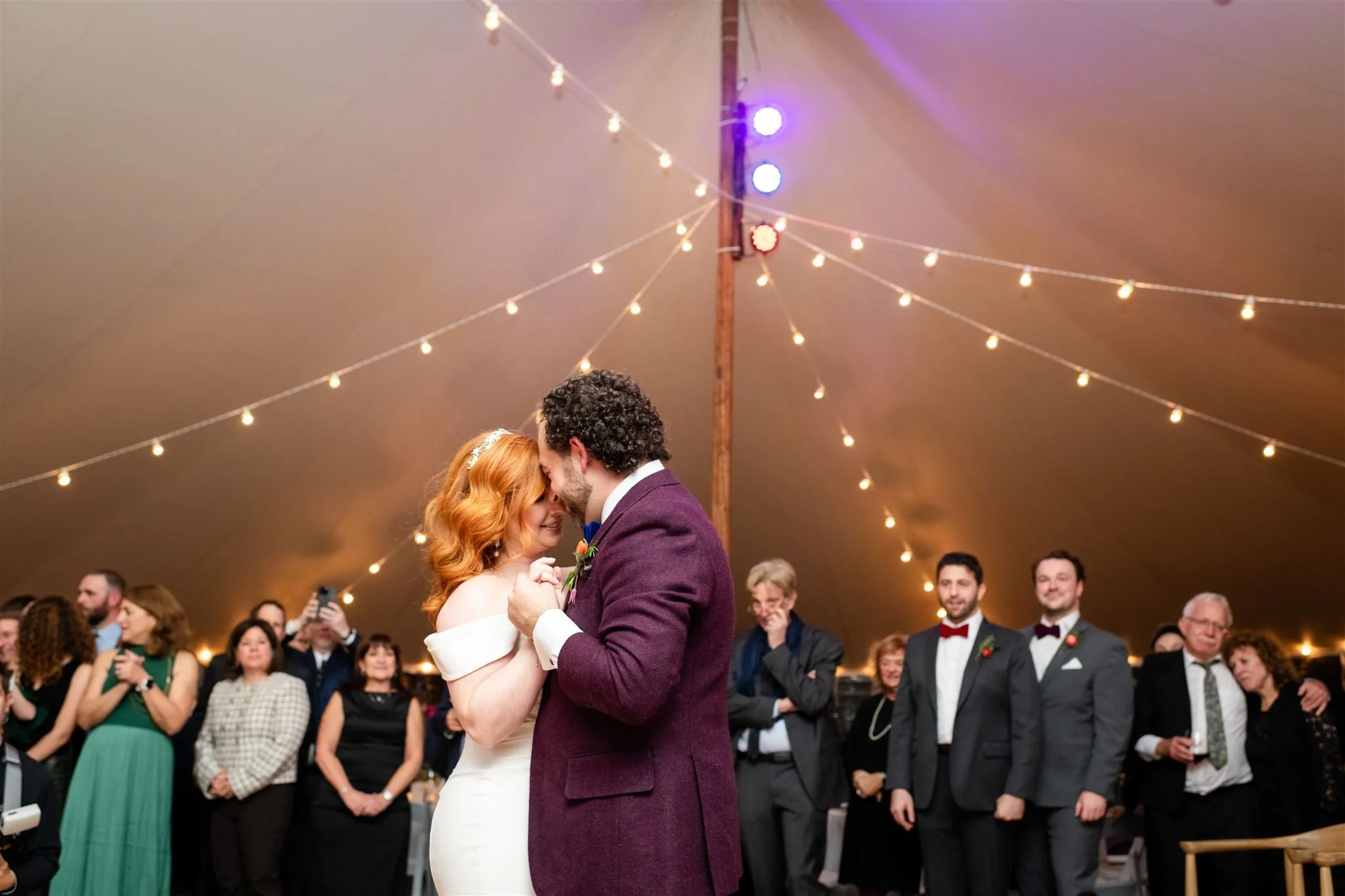 A bride and groom are dancing closely at their wedding reception under a canopy of string lights and purple lighting, with guests watching and smiling in the background during a wedding reception at Lambert's Cove Inn on Martha's Vineyard .