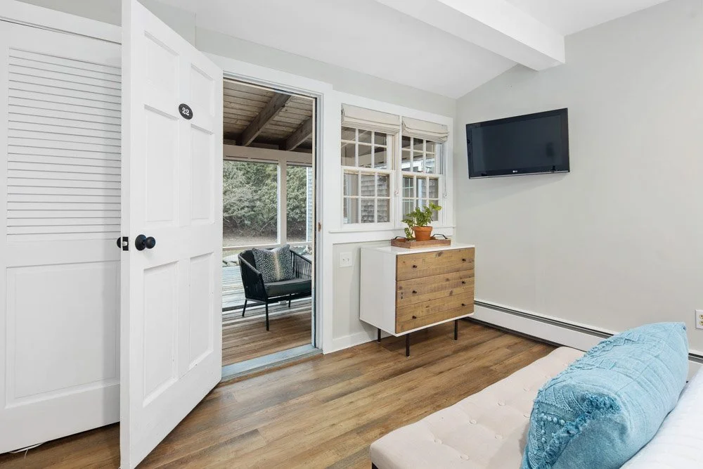 Interior of a bedroom with white walls, a wooden floor, and a door leading to a porch with outdoor seating. The room has a wall-mounted TV, a small wooden cabinet with a potted plant on top, and a bed with a light-colored headboard and blue pillows.