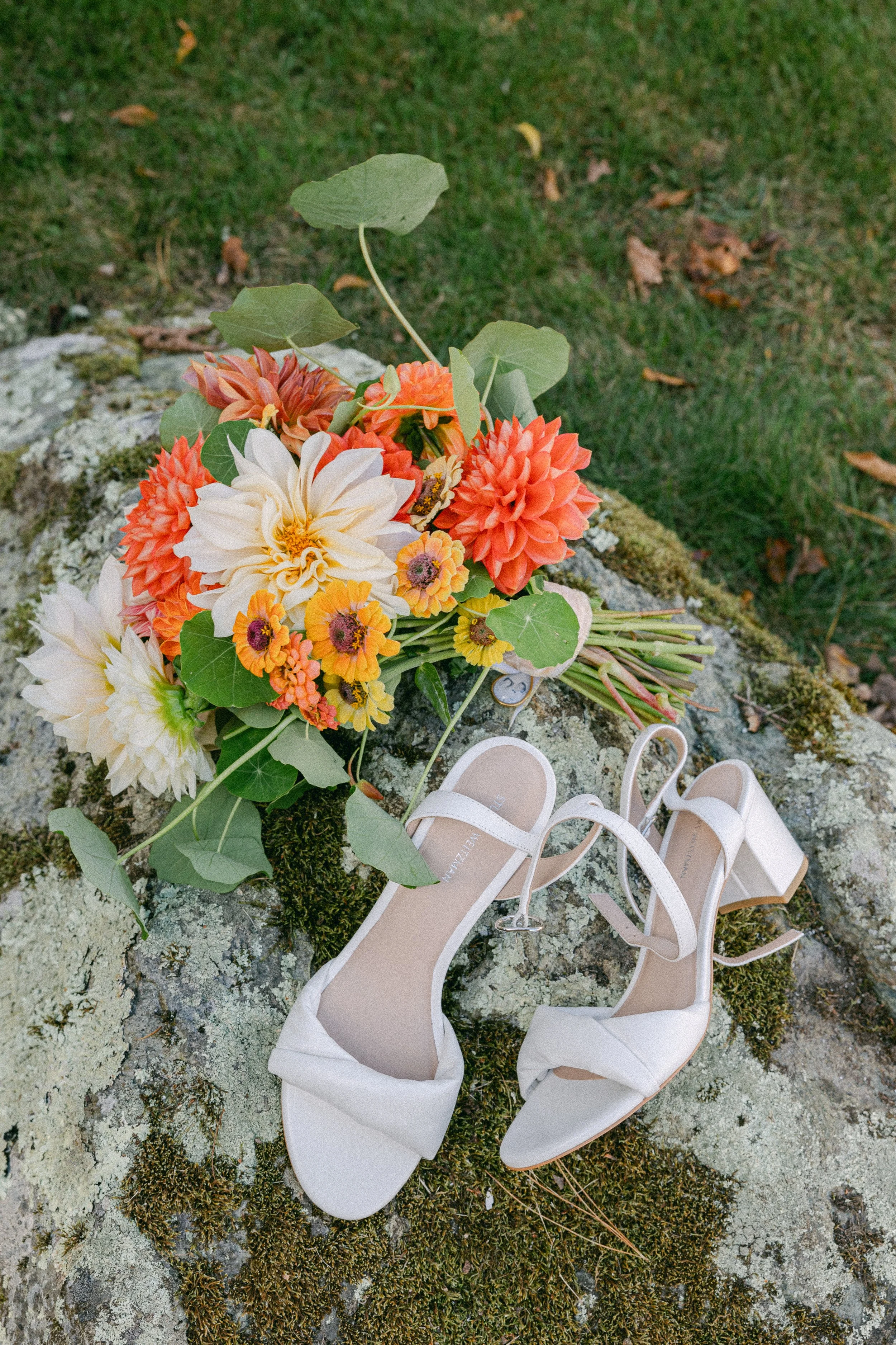 White high-heeled shoes and a bouquet of orange, white, and yellow flowers are placed on mossy, rocky ground during a wedding reception at Lambert's Cove Inn on Martha's Vineyard.