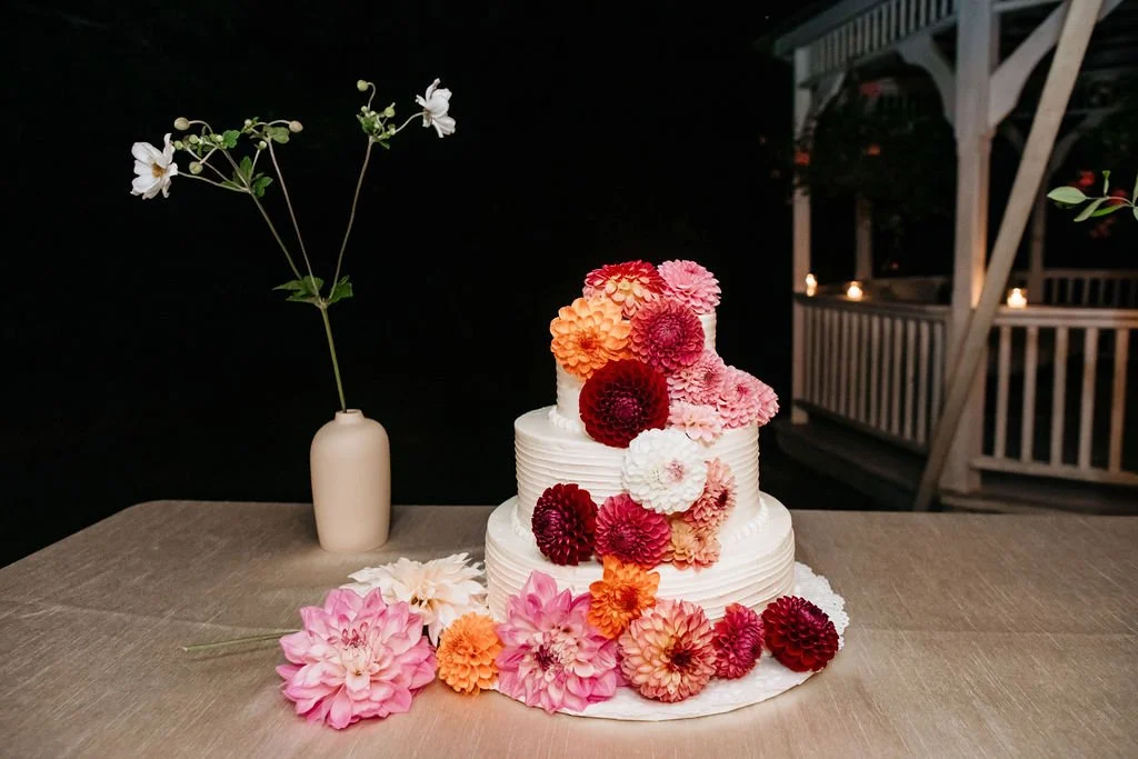 A white, two-tiered wedding cake decorated with pink, red, and orange flowers, with some flowers also placed on the table in front of the cake. There is a tall, white vase with a green-stemmed white flower on the left side of the cake. The setting ap