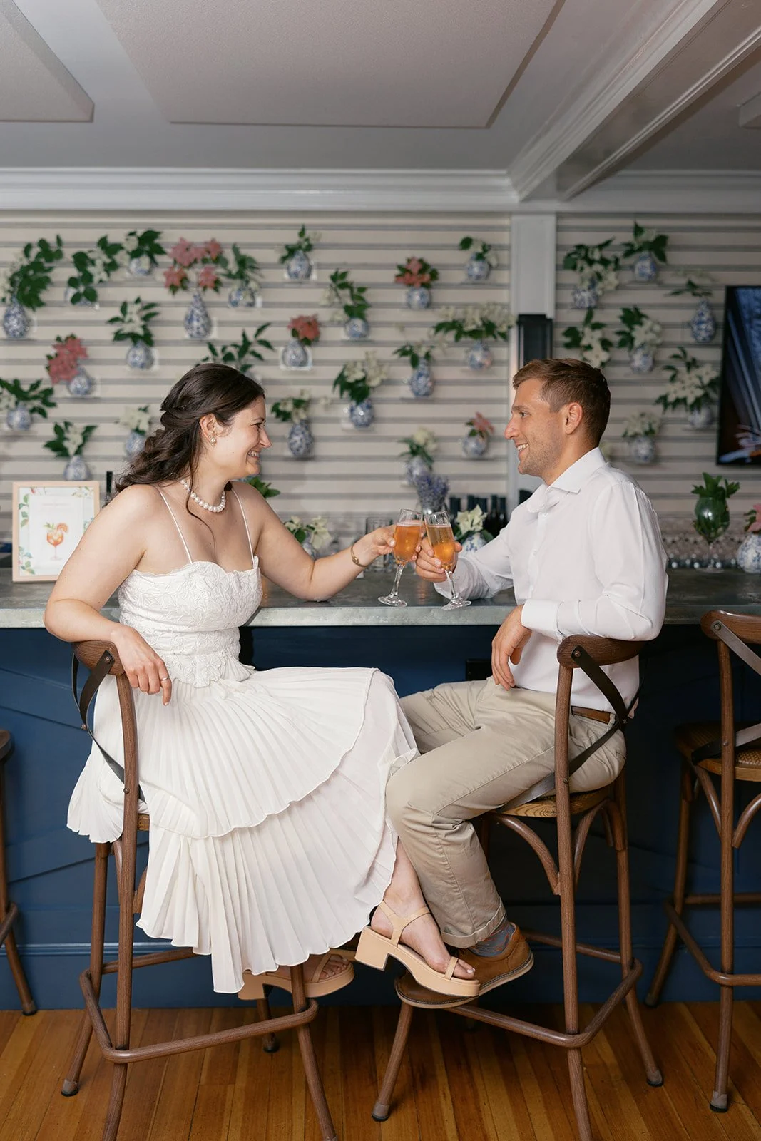A couple sitting at a bar at Lambert's Cove Inn on Martha's Vineyard, clinking glasses of champagne, smiling and looking at each other, celebrating in a decorated space with floral wall art.