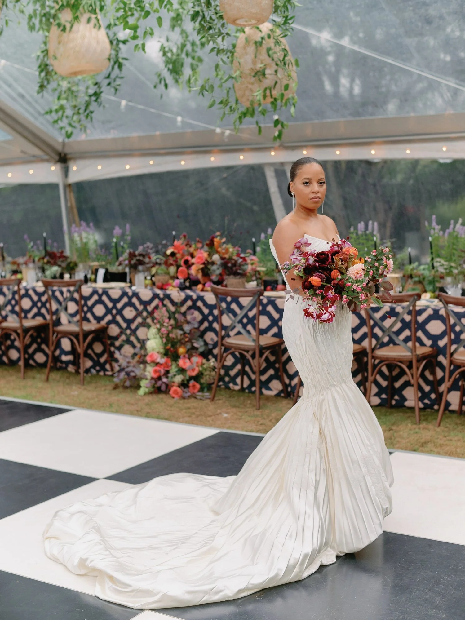 A bride in a white wedding dress holding a bouquet of pink and purple flowers at an indoor wedding reception at Lambert's Cove Inn on Martha's Vineyard with plants and decorative lighting, surrounded by tables with floral arrangements and chairs.