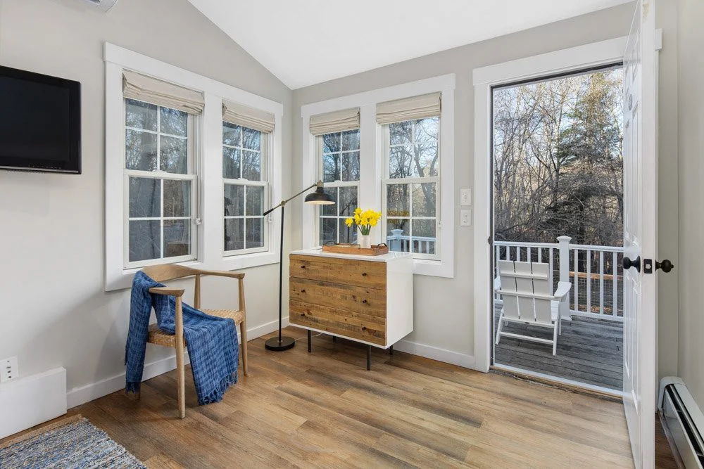Sunlit corner of a cozy room with three windows, a wooden chair with a blue blanket, a wooden cabinet with a vase of yellow flowers, and a door leading to an outdoor balcony with a white chair, overlooking trees.