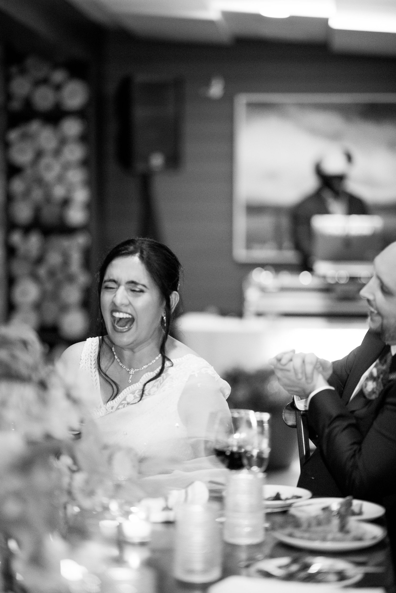 A woman with dark hair, wearing earrings and a white off-shoulder dress, is smiling and appears to be laughing during a celebration or event at a table with food and drinks. There is a man in a dark suit and white shirt holding a fork, in front of he
