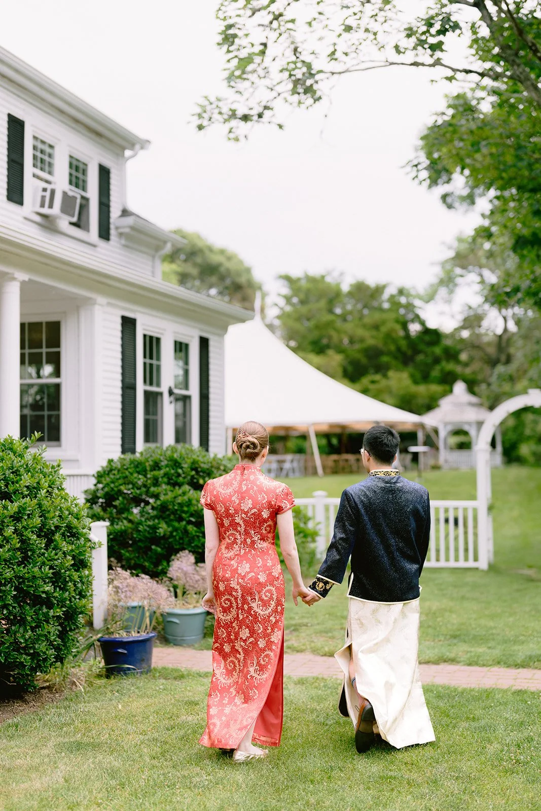 A couple wearing traditional Asian attire walking hand in hand in a garden, with a white house, a tent, and greenery in the background during a wedding at Lambert's Cove Inn on Martha's Vineyard.
