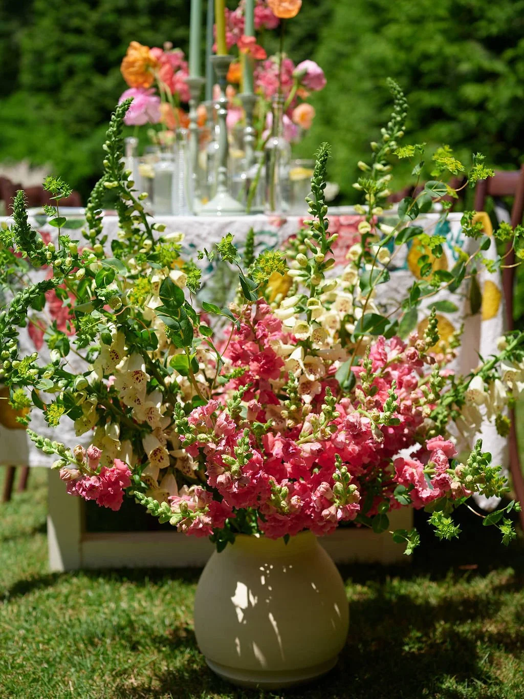 Colorful flower arrangement with pink, yellow, and white flowers in a beige vase on grass, with a decorated table in the background.