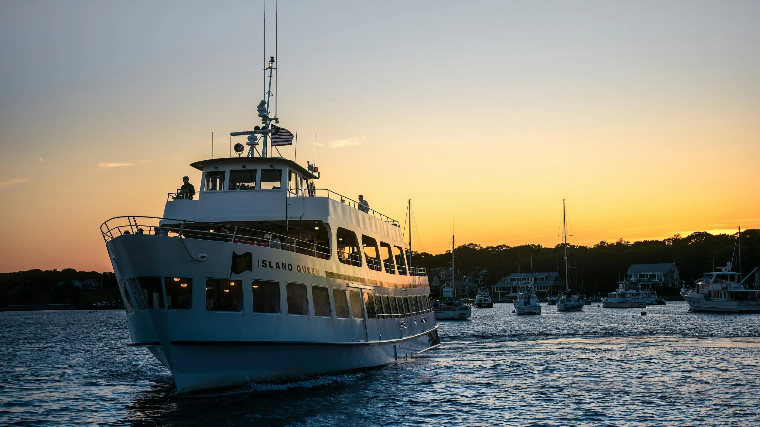 A white cruise boat named 'Island Queen' sailing on a harbor during sunset with other boats and houses in the background.