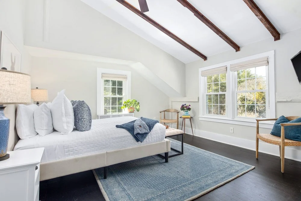 Bright bedroom with white walls, dark wood floors, a large bed with white bedding, multiple pillows, and a navy blanket. There are two windows with white blinds, a small table with a plant, and a chair with flowers. Exposed wooden beams are on the ceiling, and a mounted TV is visible.