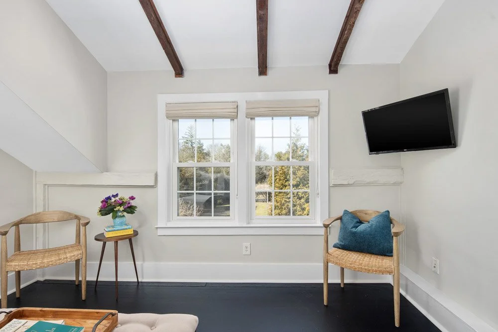 Room at Lambert's Cove Inn on Martha's Vineyard with two windows, wooden beams on ceiling, a wall-mounted flat-screen TV, two wooden chairs, a small table with a vase of flowers and books, and a wall with light paint.
