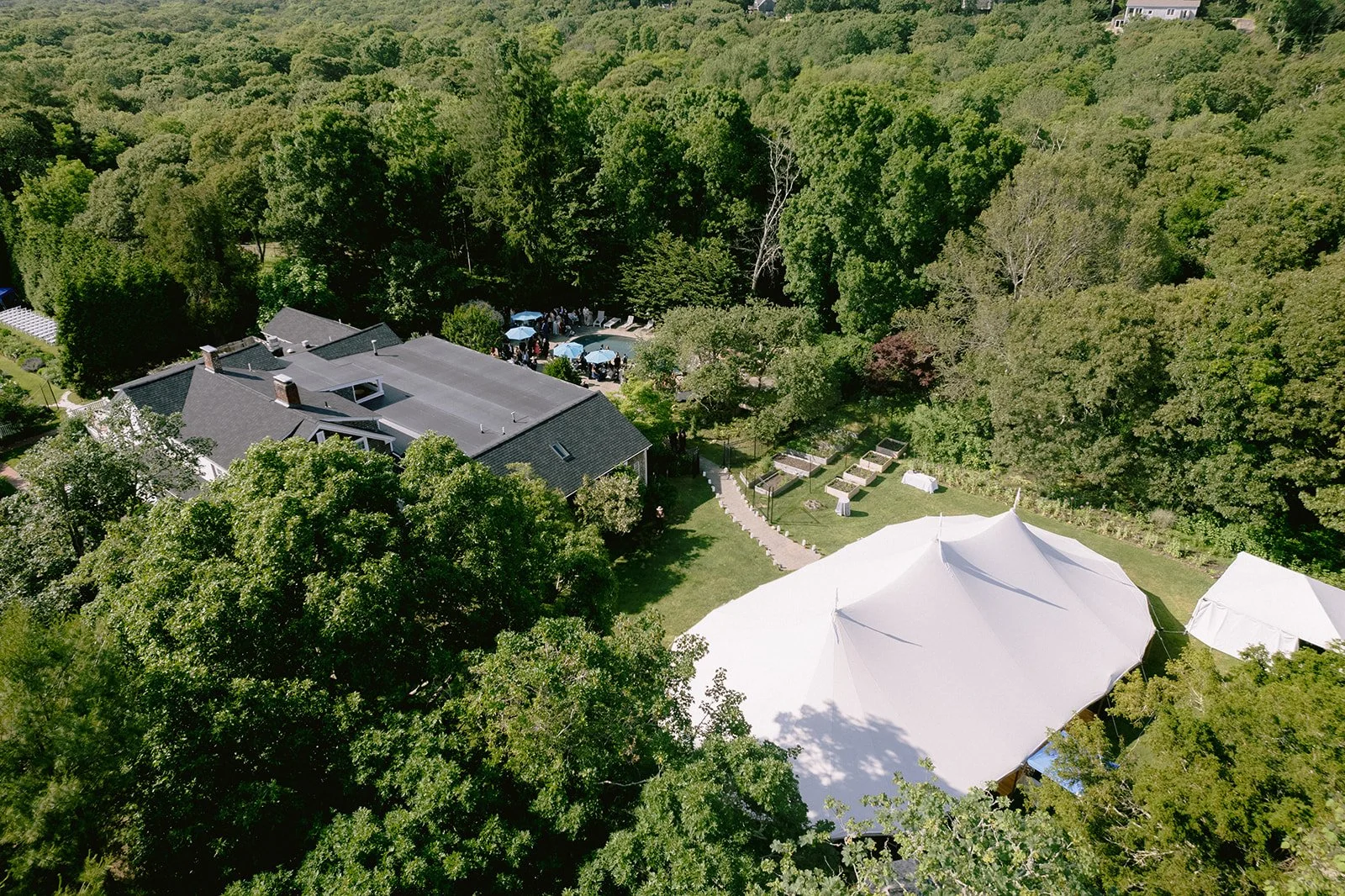 Bird's eye view the outdoor wedding venue at Lambert's Cove Inn on Martha's Vineyard, with a large white tent, smaller tents, trees, and a house with a gray roof surrounded by lush green forest.