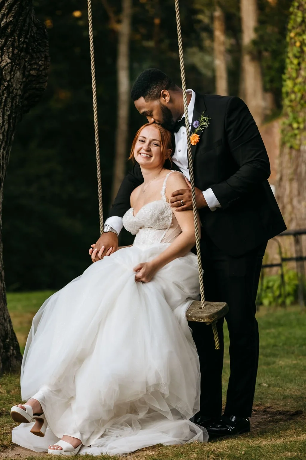 A newlywed couple in wedding attire on a wooden swing outdoors. The bride is sitting, smiling and holding her dress, while the groom is standing behind her, leaning down and kissing her forehead.