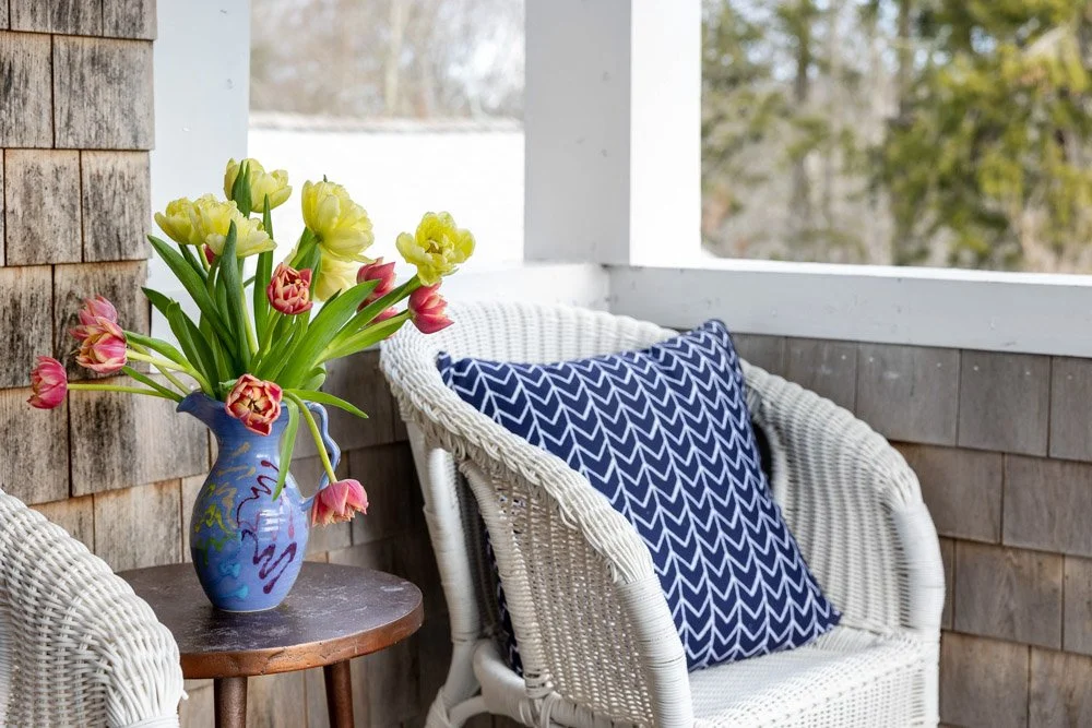 A cozy porch with a wicker chair, blue patterned pillow, wooden side table, and a vase of pink and yellow tulips.
