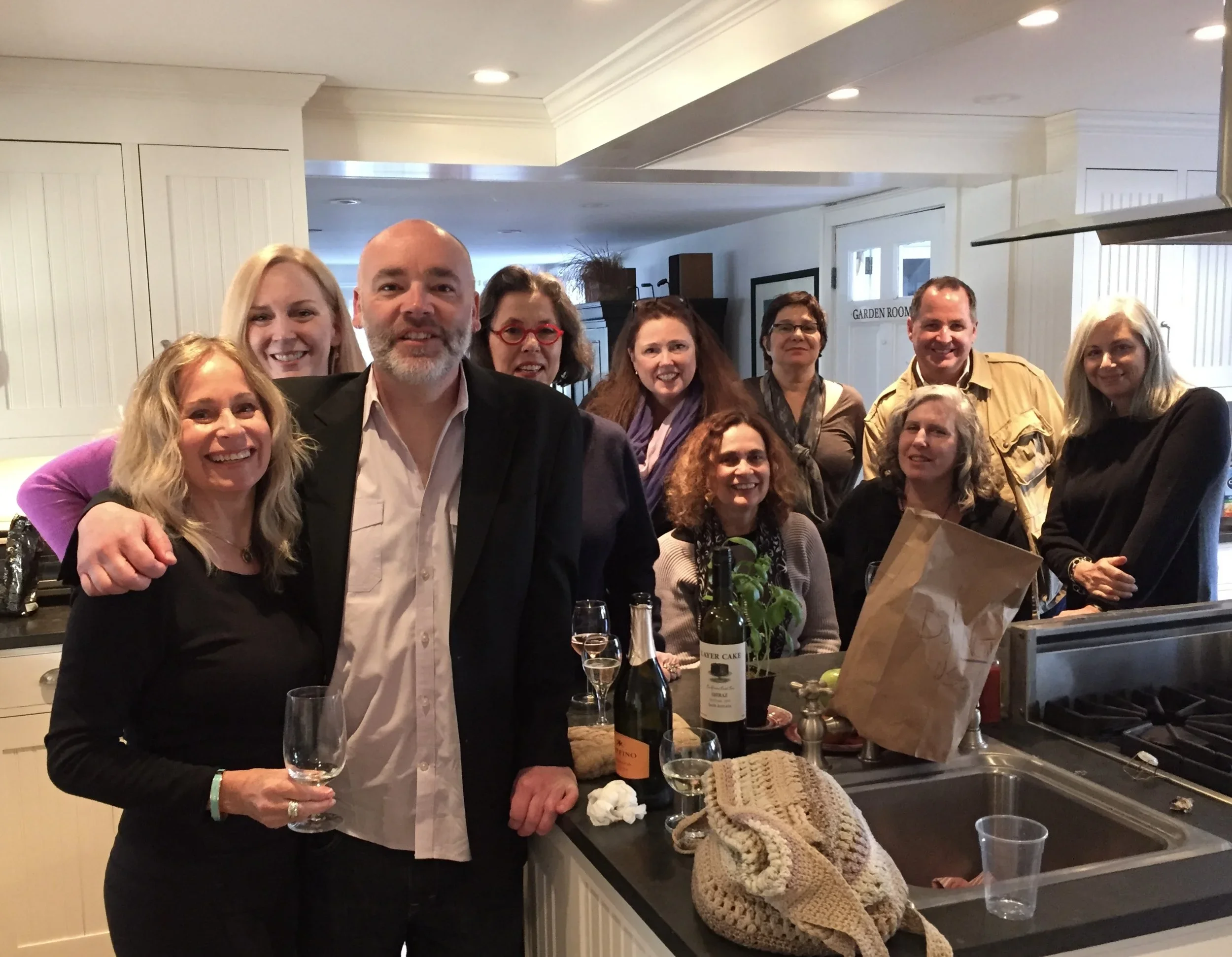 A group of people gathered in a kitchen or dining area, smiling and raising glasses for a toast. The setting appears to be a celebratory gathering, with various food and drink items on the counter, including a bottle of wine and wine glasses. The background features white cabinets, a sign that says 'Garden Room', and modern lighting.