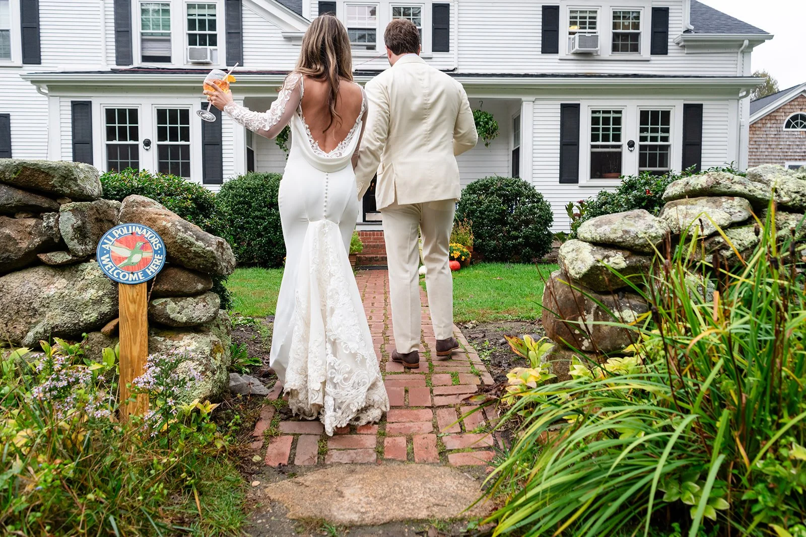 A bride and groom walk down a brick pathway toward Lambert's Cove Inn on Martha's Vineyard, holding drinks, with a sign that says 'All Polinators Welcome Here' in the front yard.