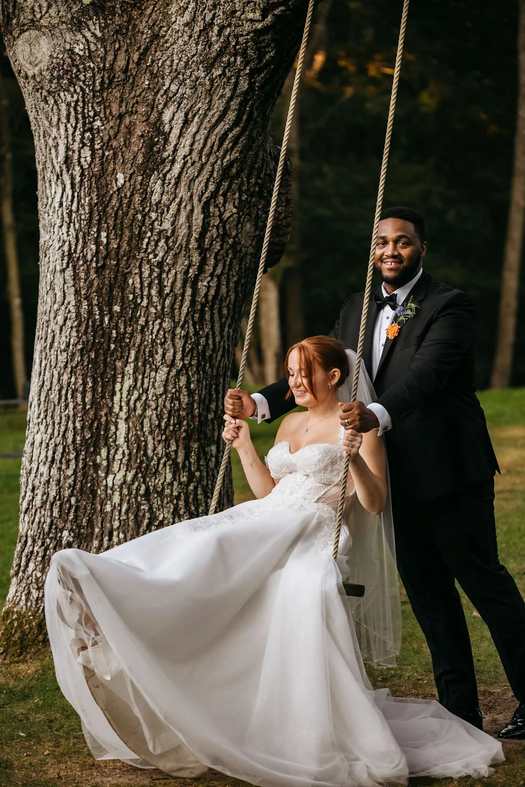 A bride with red hair in a white wedding gown sitting on a swing, being pushed by a groom in a black tuxedo, outdoors near a large tree during sunset at a wedding at Lambert's Cove Inn on Martha's Vineyard.