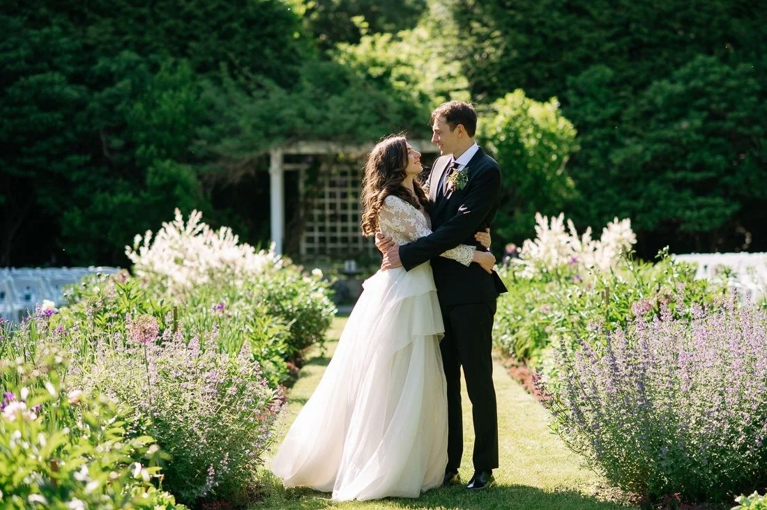 A bride and groom hugging outdoors in a garden with blooming flowers and green trees.