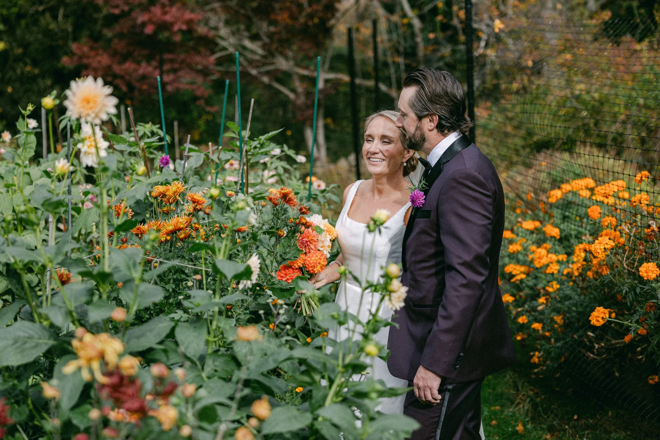 A bride and groom in wedding attire smiling and talking with each other on a flower-filled garden path surrounded by orange and pink flowers at Lambert's Cove Inn on Martha's Vineyard.