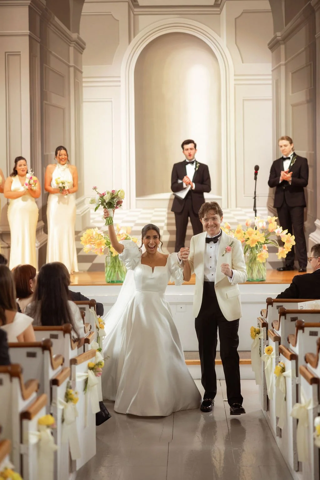 The bride and groom celebrating their wedding ceremony, smiling, holding hands, and holding flowers inside a decorated church with guests and wedding party applauding.