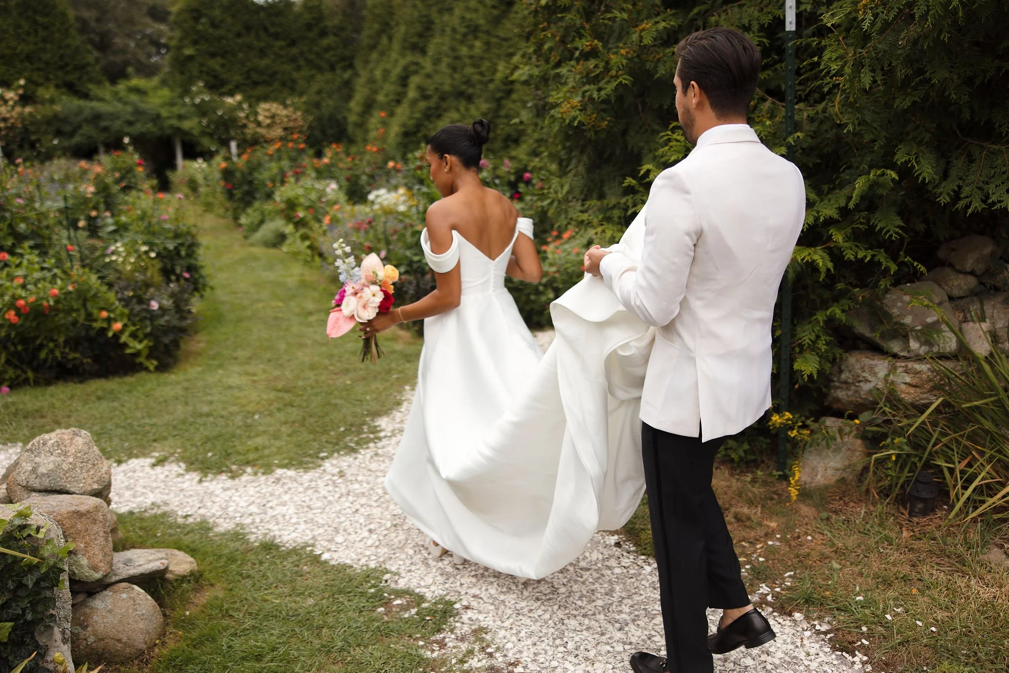 A bride and groom walking through the English garden at Lambert's Cove Inn on Martha's Vineyard, with the bride holding a bouquet of flowers and a woman in a white dress leading the way.