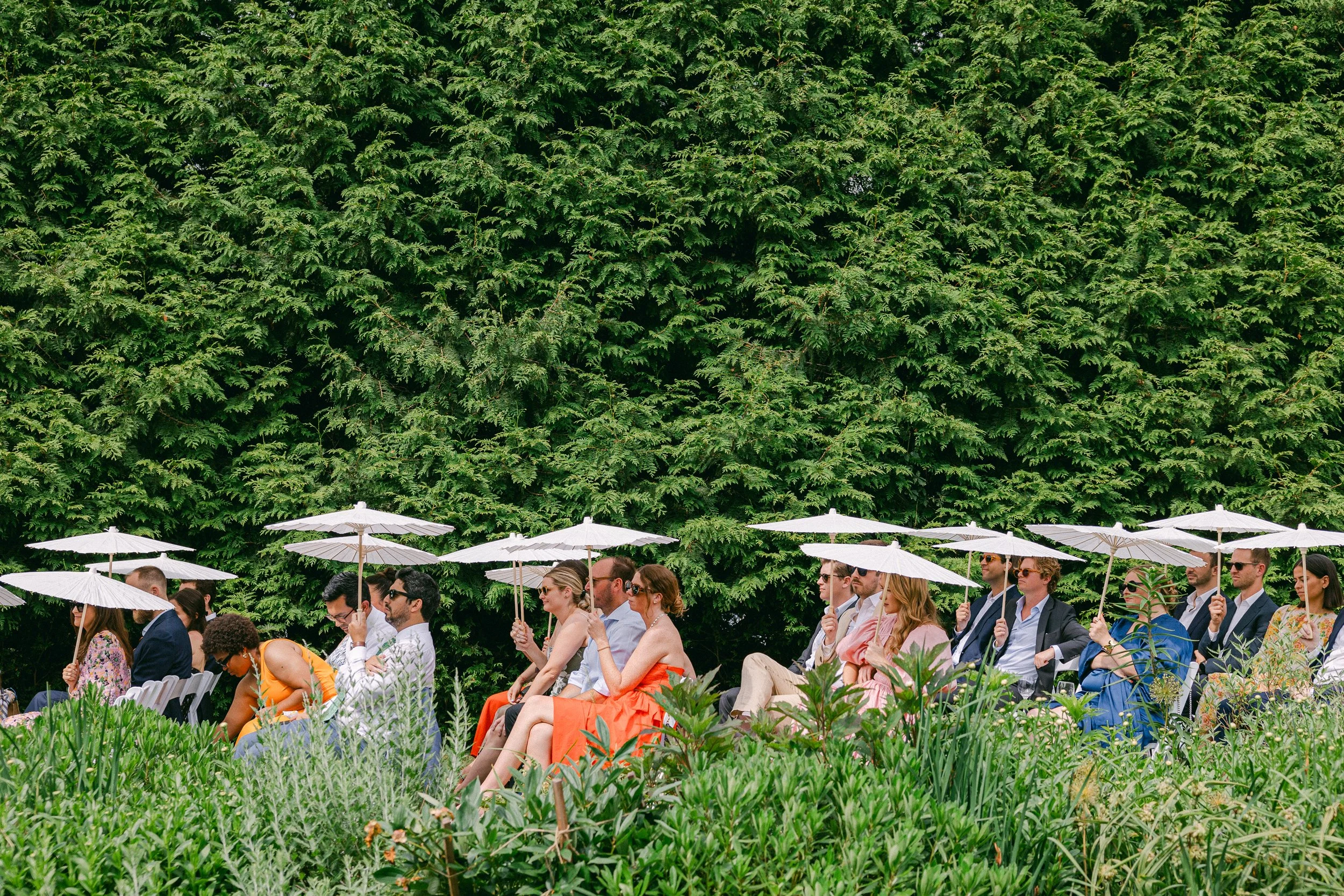 A group of people attending a wedding at Lambert's Cove Inn on Martha's Vineyard, sitting under white umbrellas with large green trees in the background.