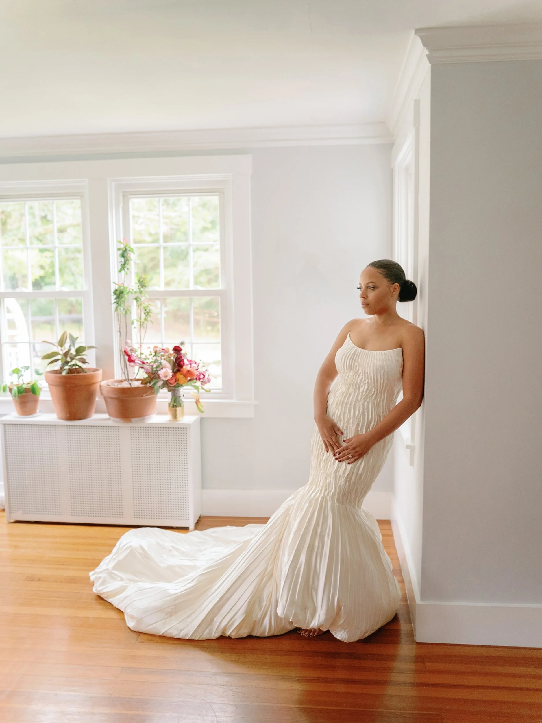 A woman in a strapless, cream-colored, textured gown with a long train stands barefoot in a bright, white-walled room with large windows, potted plants, flowers, and hardwood floors, leaning against a corner.
